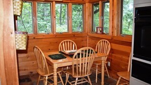 Une cabane en bois avec une table à manger et quatre chaises, de grandes fenêtres donnant sur la forêt et un four encastré.