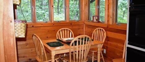 Una cabaña de madera con una mesa de comedor y cuatro sillas, ventanas amplias que ofrecen vistas al bosque y un horno empotrado.
