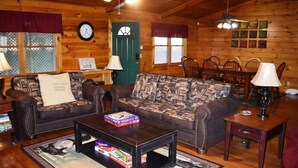 Interior acogedor de una cabaña de troncos con un sofá, dos sillones, una mesa de centro y una mesa de comedor con sillas.