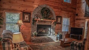A cozy log cabin interior with a stone fireplace, plaid furniture, and a television. - Mountain Rivers Lodge (Mineral Bluff)