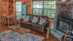 A rustic cabin interior with a stone fireplace, wooden walls, a leather sofa, a dining table with chairs, and a blue cabinet. - Squirrelled Away (Cherry Log)