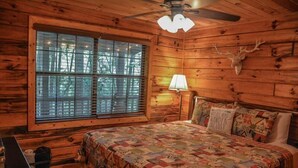 A wooden cabin bedroom with a bed, a window with blinds, a ceiling fan, and a deer head mounted on the wall. - Squirrelled Away (Cherry Log)