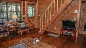 A wooden cabin interior with a staircase, a television on a stand, and a desk with a lamp and chair. - Squirrelled Away (Cherry Log)