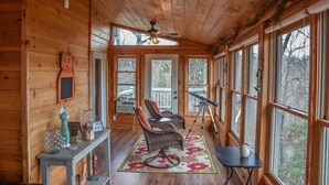 Een gezellig chaletinterieur met houten wanden, een plafondventilator, grote ramen, rieten stoelen, een kleine tafel en een vloerkleed.