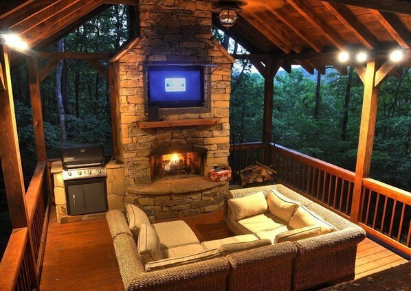 A cozy outdoor living area with a stone fireplace, a television mounted on the wall, and a comfortable seating arrangement.