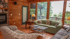 A cozy cabin interior with a fireplace, wooden walls, a green sofa, two patterned armchairs, and a dining table. - Picabo (Blue Ridge)