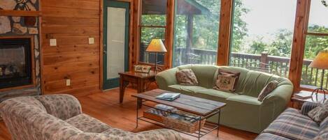 A cozy cabin interior with a fireplace, wooden walls, a green sofa, two patterned armchairs, and a dining table.