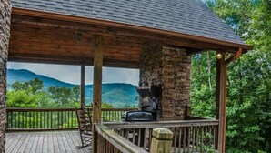 A covered outdoor area with a stone fireplace, wooden deck, and a view of green hills. - Dream Catcher (Morganton)