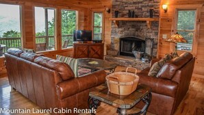 A cabin interior with a stone fireplace, leather sofas, a television, and a large window overlooking greenery. - Dream Catcher (Morganton)