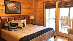 A wooden cabin bedroom with a bed, pillows, a framed picture, a lamp, and a sliding glass door leading to an outdoor area with a chair. - Mi Casa es su Casa (Blue Ridge)