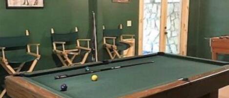 A pool table with a green felt surface, surrounded by green walls and wooden chairs.