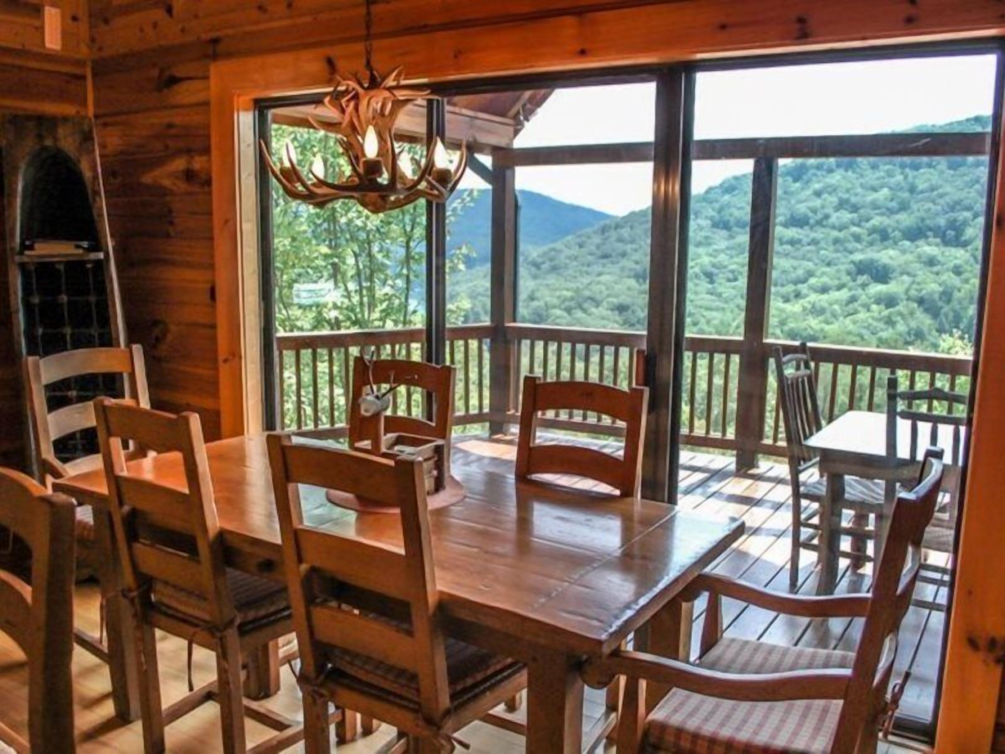 A wooden cabin with a dining area featuring a large table and chairs, a chandelier, and a balcony with a scenic mountain view.