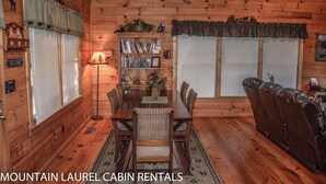Interior de cabana com mesa de jantar, cadeiras, sofá, estante e janela com persianas.