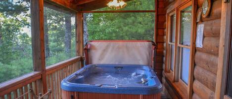 A wooden cabin with a hot tub on the porch, wooden railings, and a view of the forest.