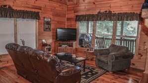 Interior acolhedor de uma cabana com um sofá de couro, poltrona e uma televisão sobre uma mesa de madeira.