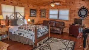 Une chambre dans un chalet en rondins, avec un lit, un canapé en cuir, une commode en bois, une horloge et un ventilateur de plafond.