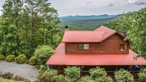 Une cabane en rondins à deux étages, surmontée d’un toit en métal rouge, entourée d’une forêt dense et avec des montagnes en arrière-plan.