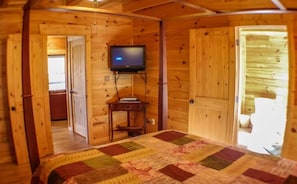 A cozy cabin room with a quilted bedspread, a television mounted on the wall, and wooden walls and doors. - The Great Escape (Morganton)