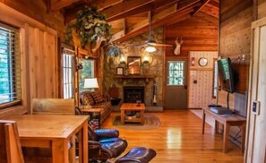 A cozy cabin interior with a wooden floor, a fireplace, a sofa, a dining table, and a television. - Vista (Blue Ridge)