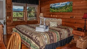 A wooden cabin bedroom with a large bed, a window with a view of mountains, and a painting of a forest. - Okeana (Blue Ridge)