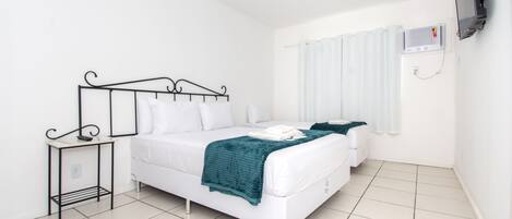 A clean, white room with a bed, a bedside table, a TV mounted on the wall, and a window with curtains.