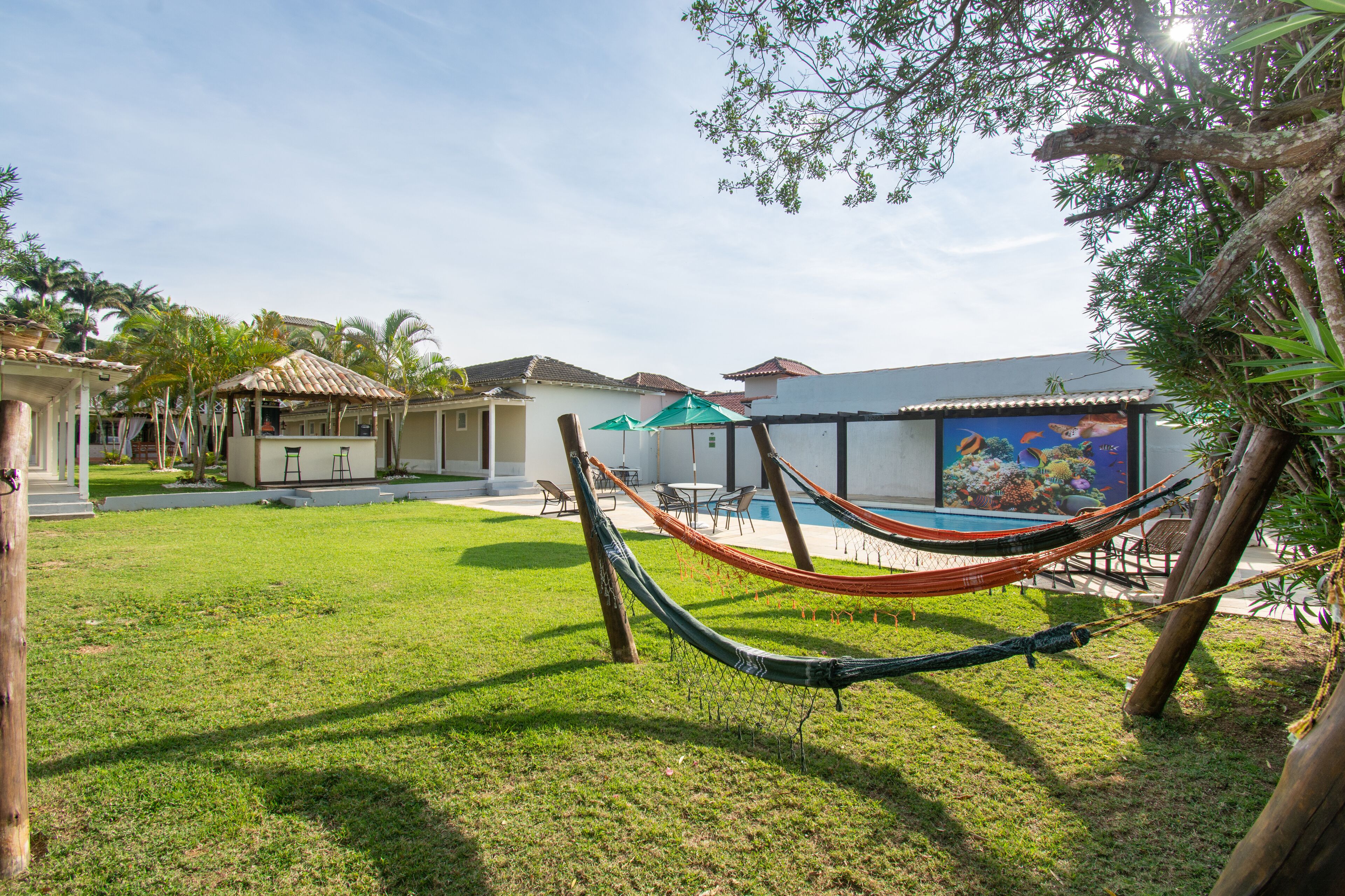 A hammock in a grassy area with a pool and a building with a tiled roof in the background.