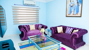 Een woonkamer met twee paarse banken, een glazen salontafel, een kleurrijk vloerkleed, een schilderij aan de muur en een airconditioner.