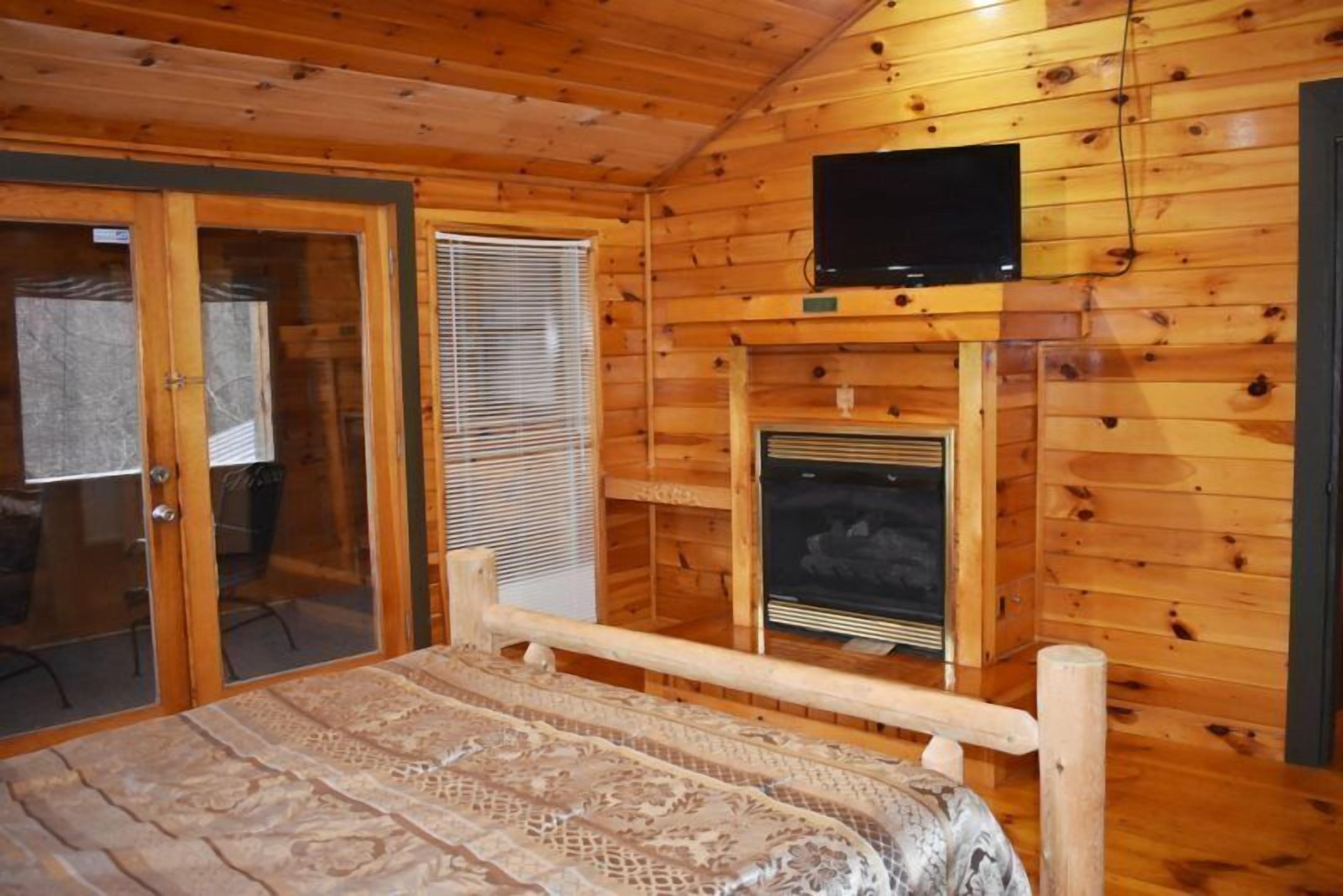 A wooden cabin room with a bed, a television mounted above a fireplace, and a sliding glass door leading outside.