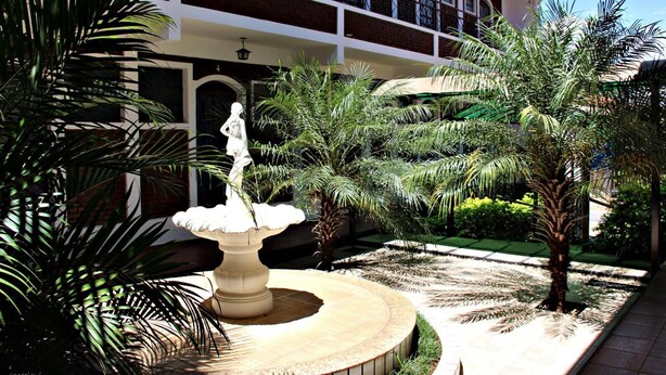 A courtyard with a central fountain, palm trees, and a building with a balcony in the background. - Hotel JS (Alfenas)