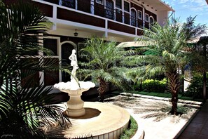 A courtyard with a central fountain, palm trees, and a building with a balcony in the background. - Hotel JS (Alfenas)
