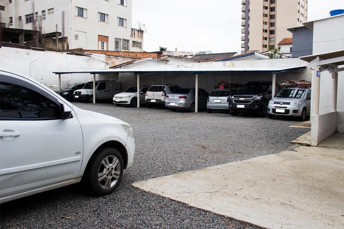 Uma área de estacionamento com vários carros, uma estrutura coberta e edifícios ao fundo.