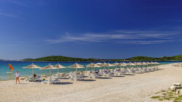 A beach with sun loungers and umbrellas, a clear sky, and a distant green hill. - Olive Garden Camp Resort (Sibenik)