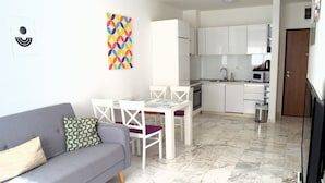 A modern living room with a grey sofa, a white dining table with chairs, a kitchen area with white cabinets, and a marble floor. - Yeshill Apartments (Budva)