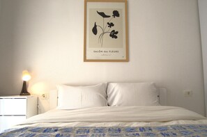Ein ordentlich bezogenes Bett mit weißen Leinen und einer blauen Bettdecke mit Muster, ein Nachttisch mit Lampe sowie ein botanisches Bild in einem Rahmen an der Wand.