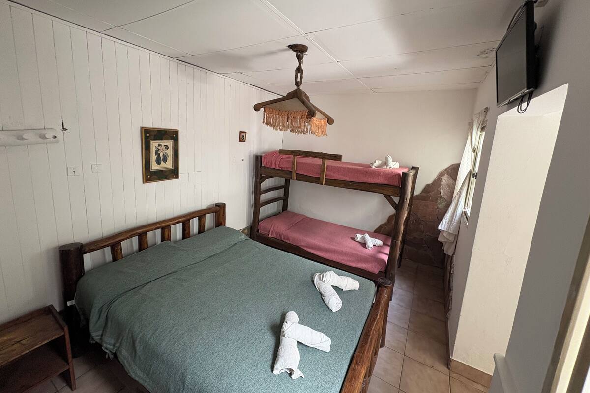 Ein Zimmer mit einem Etagenbett, einem Einzelbett, einer hölzernen Bank, einem Deckenventilator, einem gerahmten Bild und einem Fernseher an der Wand.