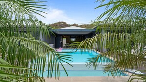 A swimming pool surrounded by palm trees, with a covered area and a view of hills in the background.