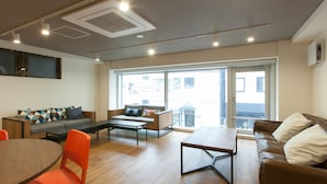 A modern living room with a wooden floor, a brown leather sofa, a grey sofa with colorful cushions, a wooden dining table with orange chairs, and a large window with a view of buildings.