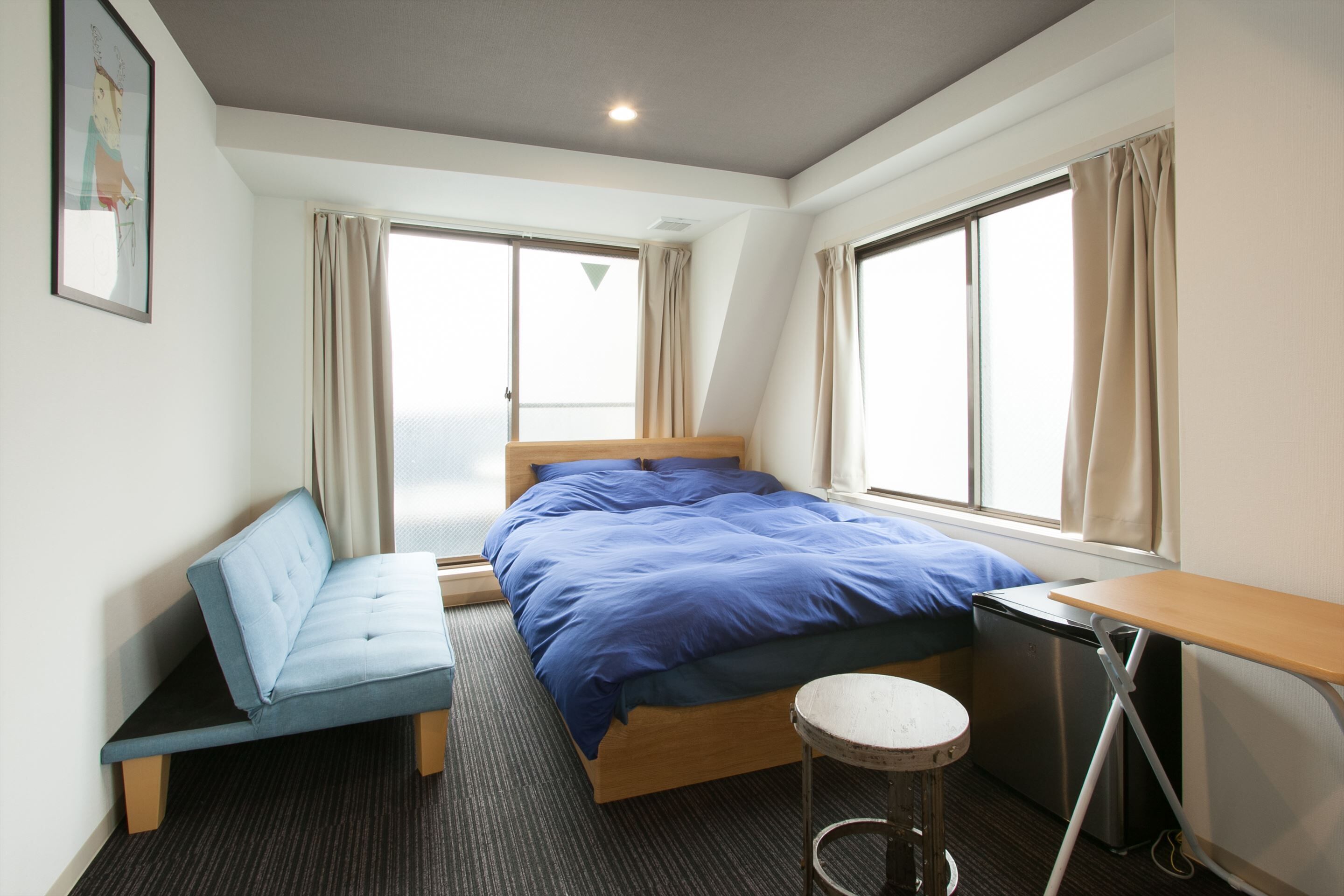 A modern hotel room with a bed, a desk, a chair, and a small stool.