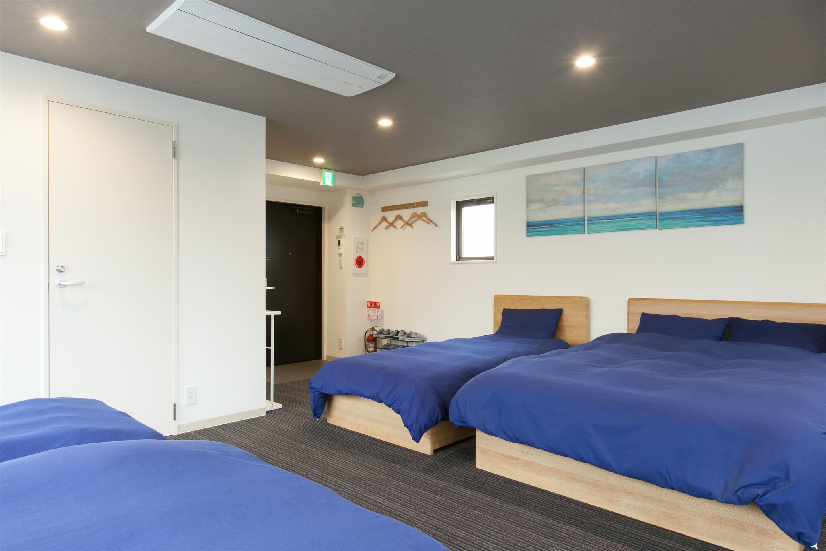 A hotel room with two beds, a painting of the ocean, a fire extinguisher, and a door to the hallway.