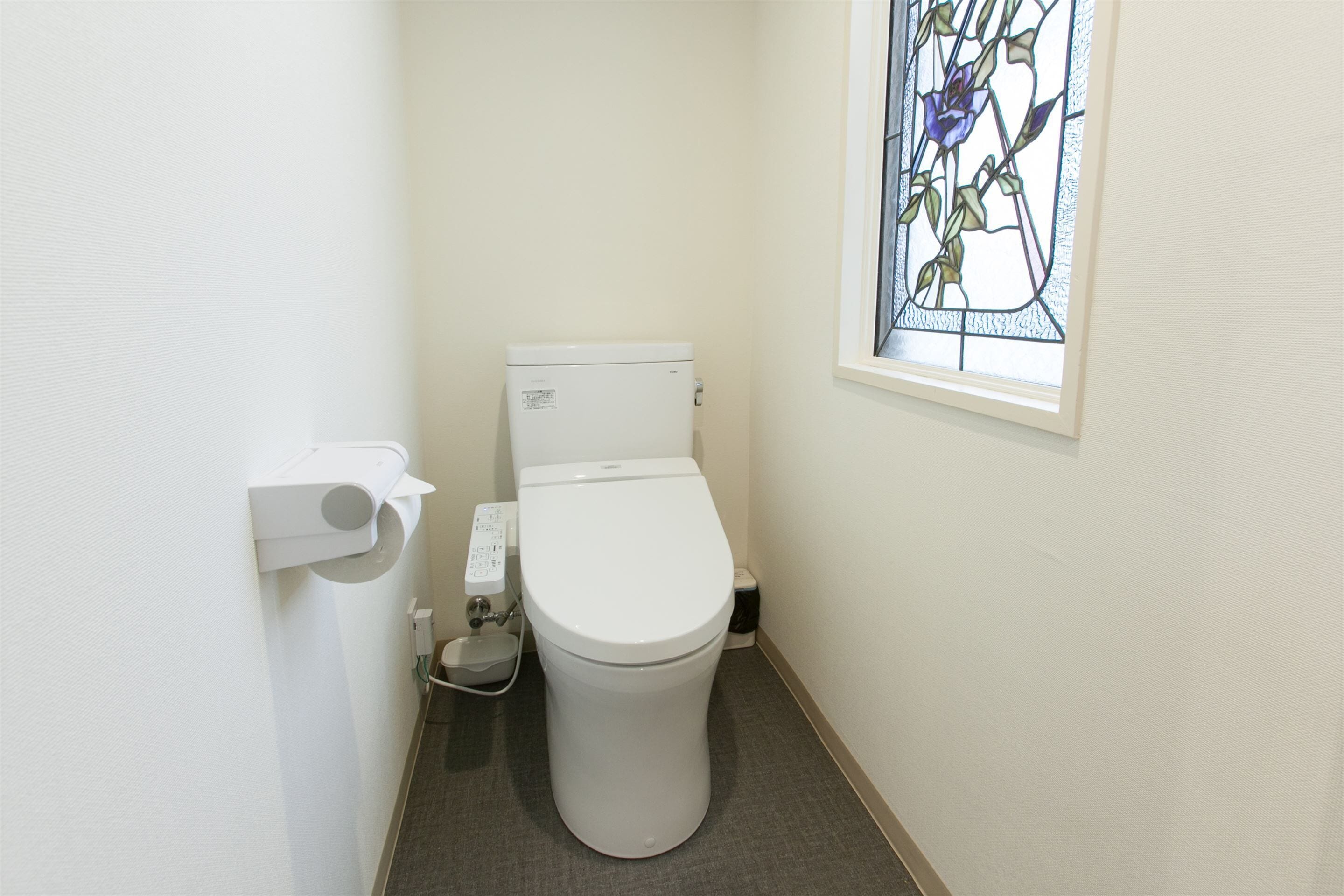 A small, clean bathroom with a toilet, a paper towel dispenser, and a stained glass window.