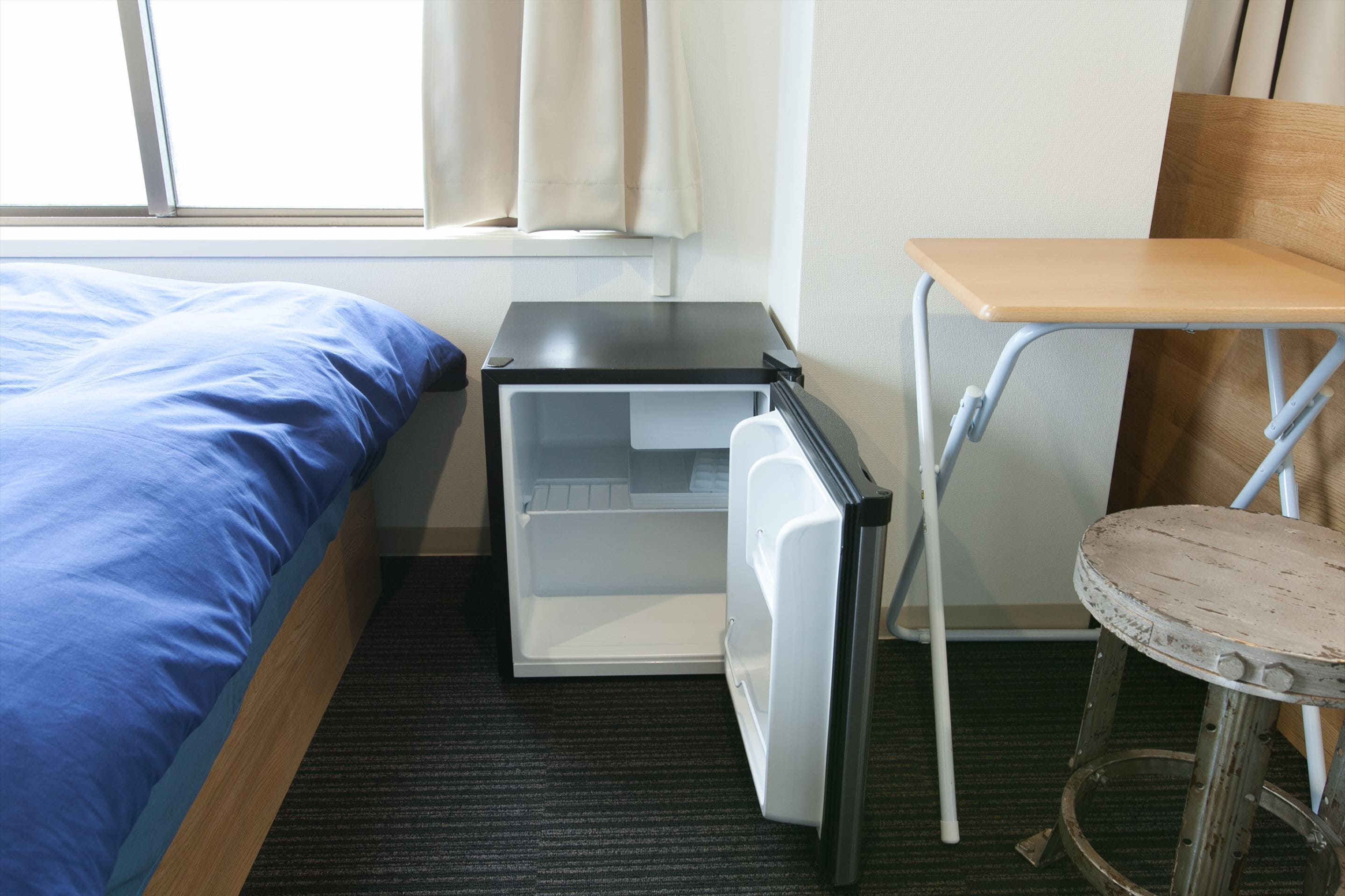 A hotel room with a bed, a small table, a stool, and a refrigerator.