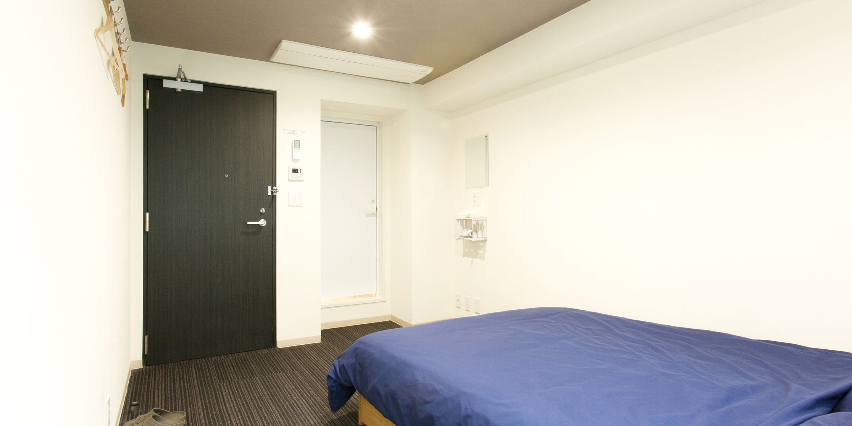 A hotel room with a bed, a door, a light switch, and a wall-mounted light fixture.
