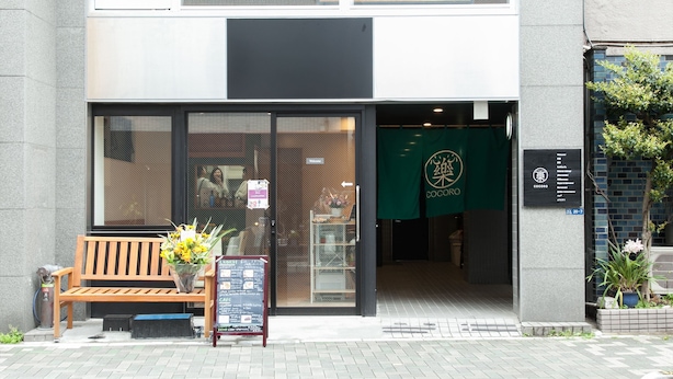 A building entrance with a wooden bench, a flower arrangement, and a green awning with a logo.