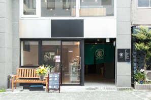 A building entrance with a wooden bench, a flower arrangement, and a green awning with a logo. - 44824 Hotel Cocoro Inn Asakusa Kuramae (Tokyo)