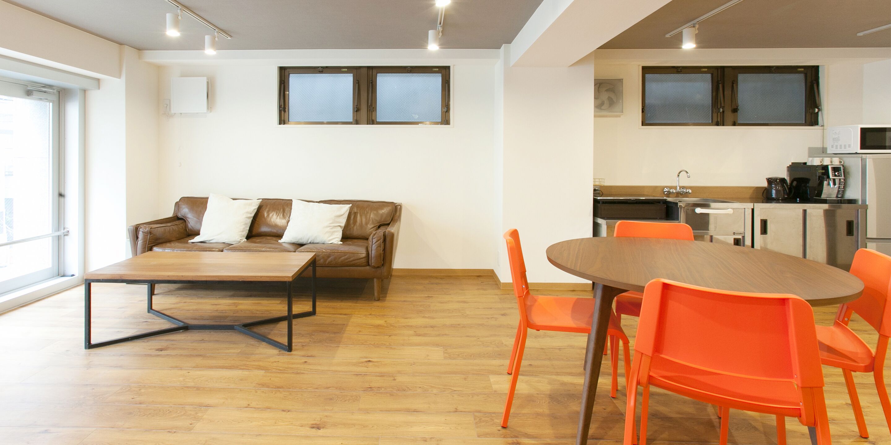Un salon moderne avec un canapé en cuir marron, une table basse en bois et des fauteuils orange. Il y a également un coin cuisine équipé d’un évier, d’une cuisinière et d’un four à micro-ondes.