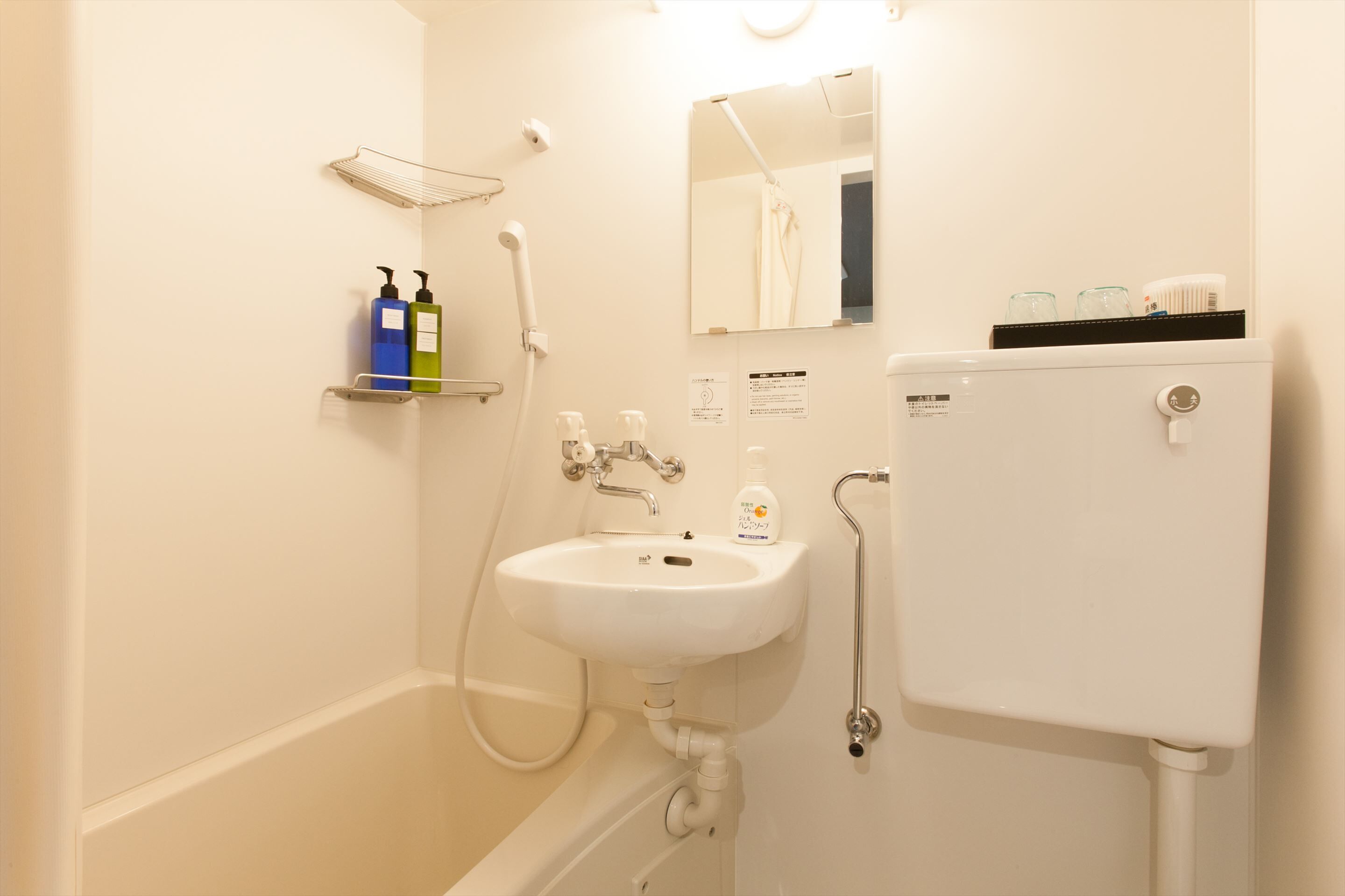 A bathroom with a white sink, a toilet, a shower, and a mirror. There are toiletries on a shelf and a sign on the wall.
