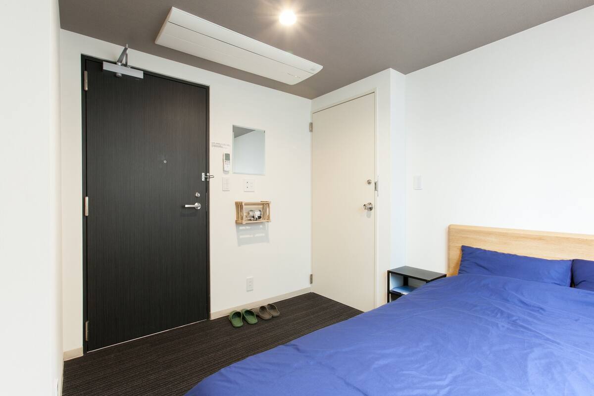 A hotel room with a bed, a wardrobe, a mirror, a small shelf, and a pair of slippers on the floor.