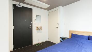 A hotel room with a bed, a wardrobe, a mirror, a small shelf, and a pair of slippers on the floor.