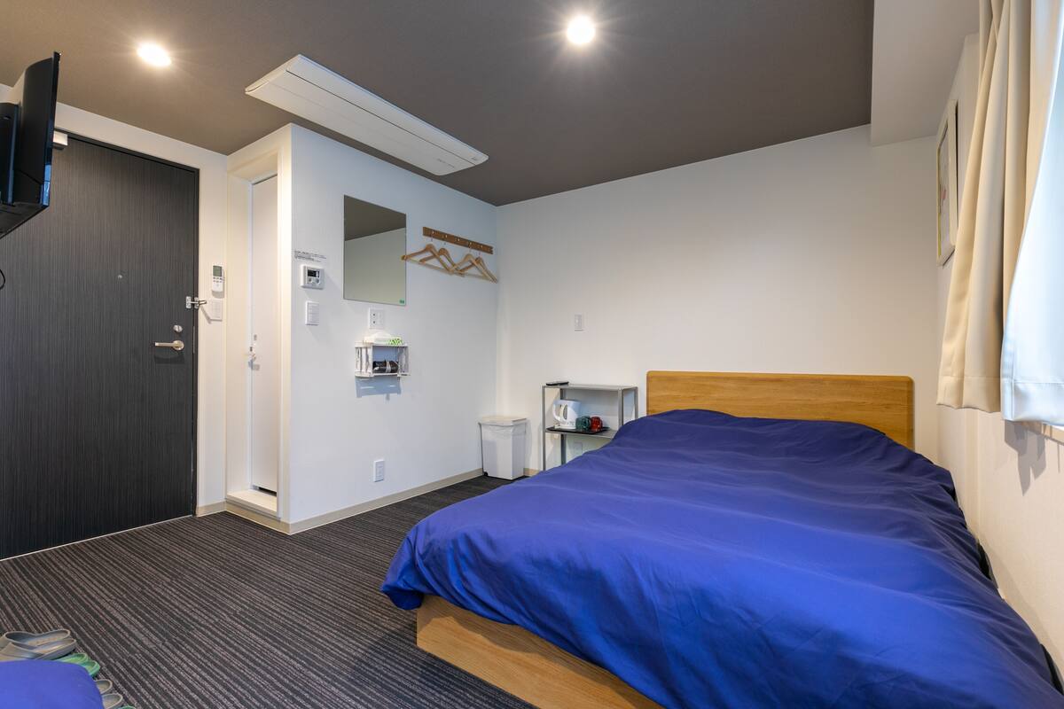 A hotel room with a bed, a TV, a mirror, a coat rack, and a small table with a kettle.