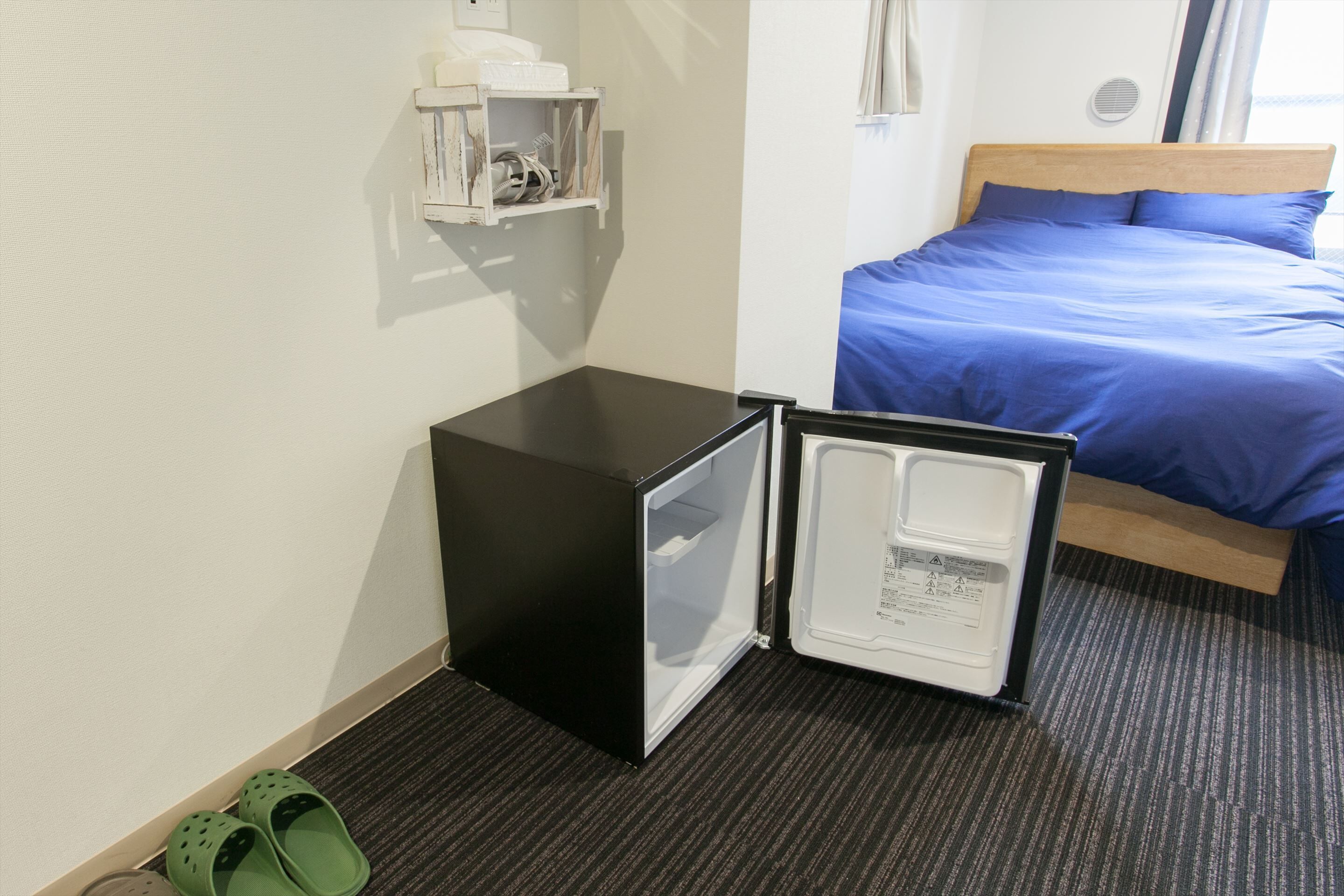 A hotel room with a bed, a small black refrigerator, and a pair of green slippers on the floor.